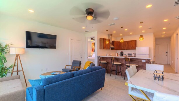 A modern living room with a blue sofa, a dining table with chairs, a kitchen area with wooden cabinets, and a ceiling fan. - Pineapple Paradise (South Padre Island)