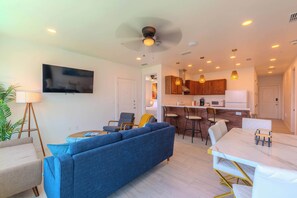 Una sala de estar moderna con un sofá azul, una mesa de comedor con sillas, una zona de cocina con armarios de madera y un ventilador de techo.