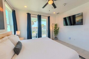 A hotel room with a large bed, a ceiling fan, a TV mounted on the wall, and a balcony with curtains.