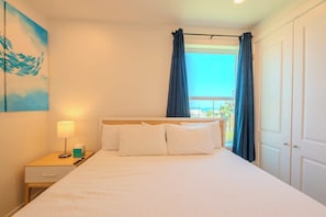 A hotel room with a bed, a nightstand, a lamp, a painting, and a view of the outdoors.