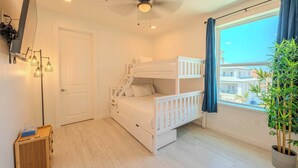 A room with a bunk bed, a ceiling fan, a window with curtains, a plant, and a small wooden table. - Pineapple Paradise (South Padre Island)