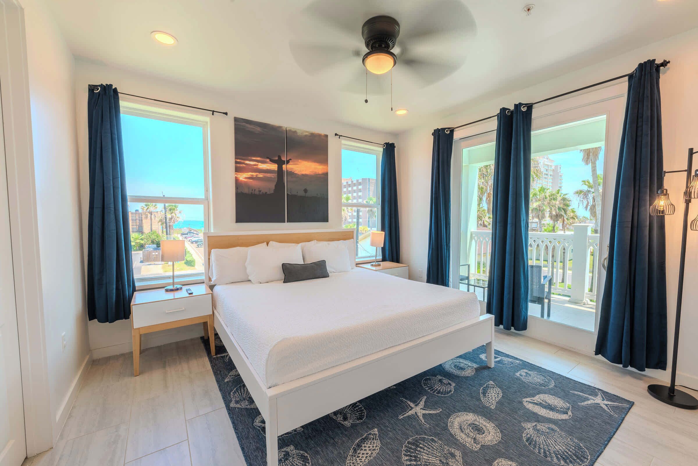 A hotel room with a bed, a nightstand, a lamp, a ceiling fan, and a view of the outside.