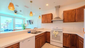 A modern kitchen with wooden cabinets, a white countertop, a built-in dishwasher, a stove, and pendant lighting. - Pineapple Paradise (South Padre Island)