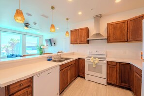 A modern kitchen with wooden cabinets, a white countertop, a built-in dishwasher, a stove, and pendant lighting. - Pineapple Paradise (South Padre Island)