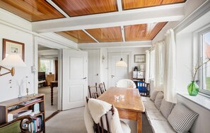 A dining area with a wooden table, chairs, and a bench. There is a bookshelf with books and a lamp. A window with a view of greenery is visible. - Stunning home in Ribe (Ribe)