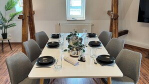 Ruokailuhuone, jossa on valkoinen pöytä, johon on katettu kuudelle, ja pöydän ympärillä on harmaita tuoleja. Katossa on puisia palkkeja, huoneessa on ruukkukasvi ja seinälle on asennettu televisio.