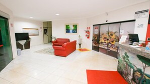 A reception area with a red sofa, a reception desk, a computer, and a painting on the wall. - HOTEL CANCIONEIRO (Sinop)