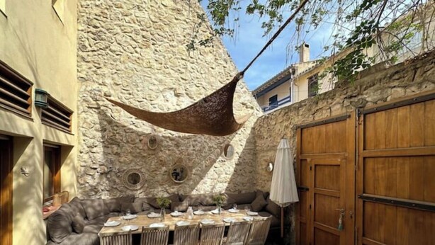 A stone-walled outdoor dining area with a wooden table and chairs, a cushioned bench, and a wooden door. - Maison Familiale Leucate Village Avec Extérieur (Leucate)