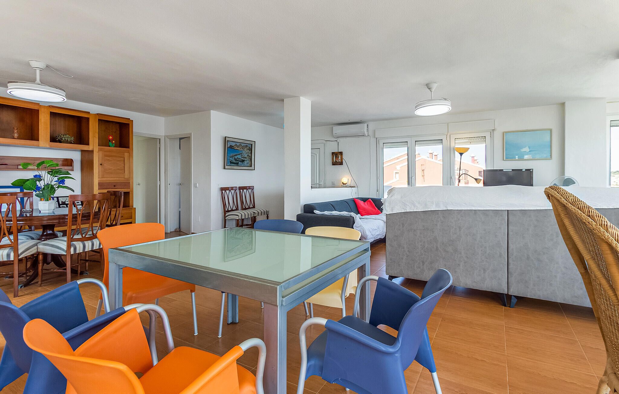 A modern living room with a dining area, featuring a glass-top table, colorful chairs, a sofa, and a large window with a view.