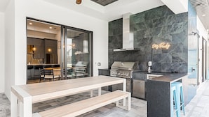 An outdoor kitchen with a grill, bar stools, and a wooden table. - Peach Paradise: Luxury Saltwater Pool & Game Room (Cape Coral)
