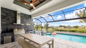 An outdoor kitchen with a grill, a table, and a view of a swimming pool. - Peach Paradise: Luxury Saltwater Pool & Game Room (Cape Coral)