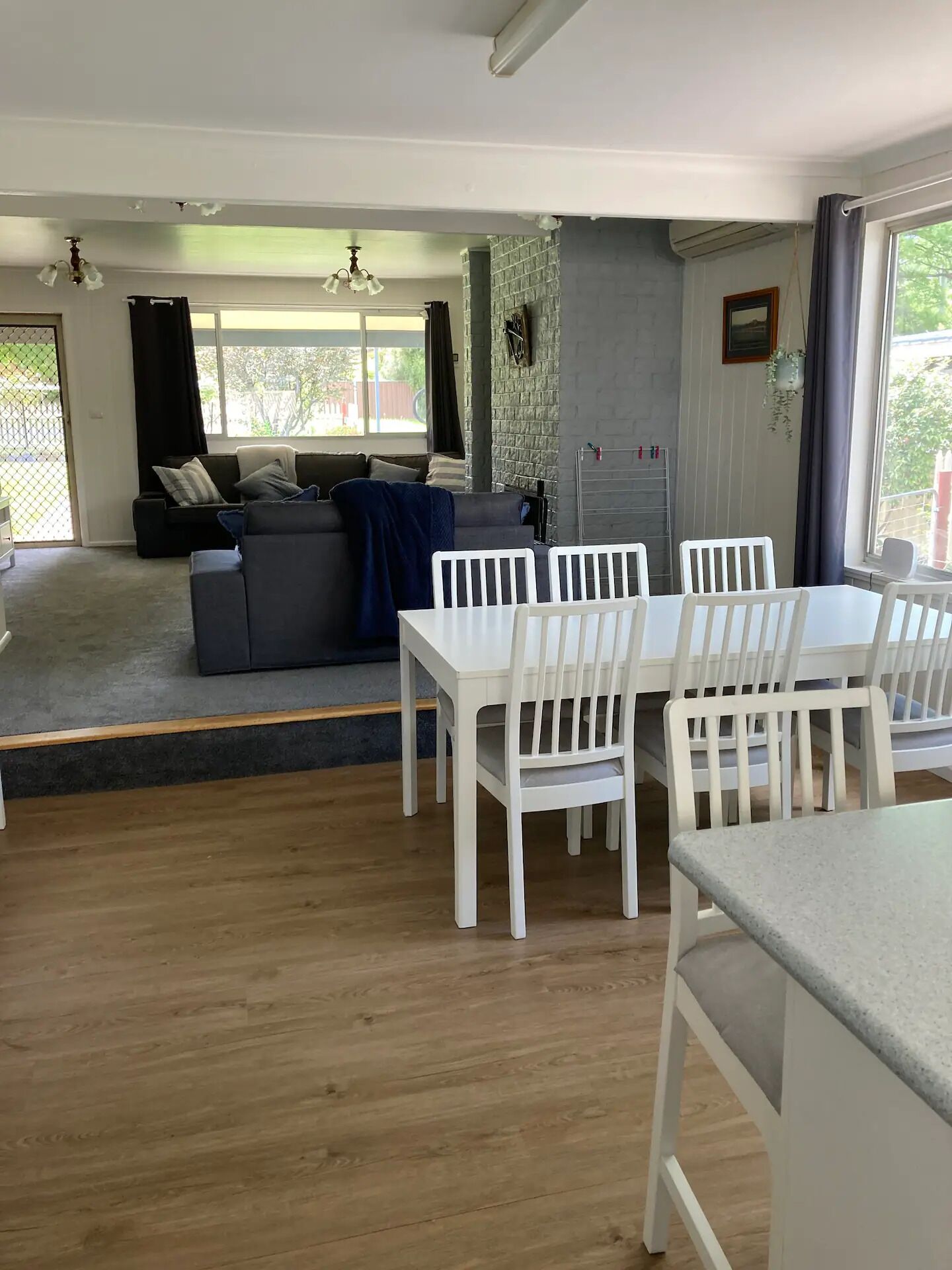 Un salon moderne avec un canapé gris, une table à manger avec des chaises blanches et un coin cuisine doté d’un plan de travail gris.