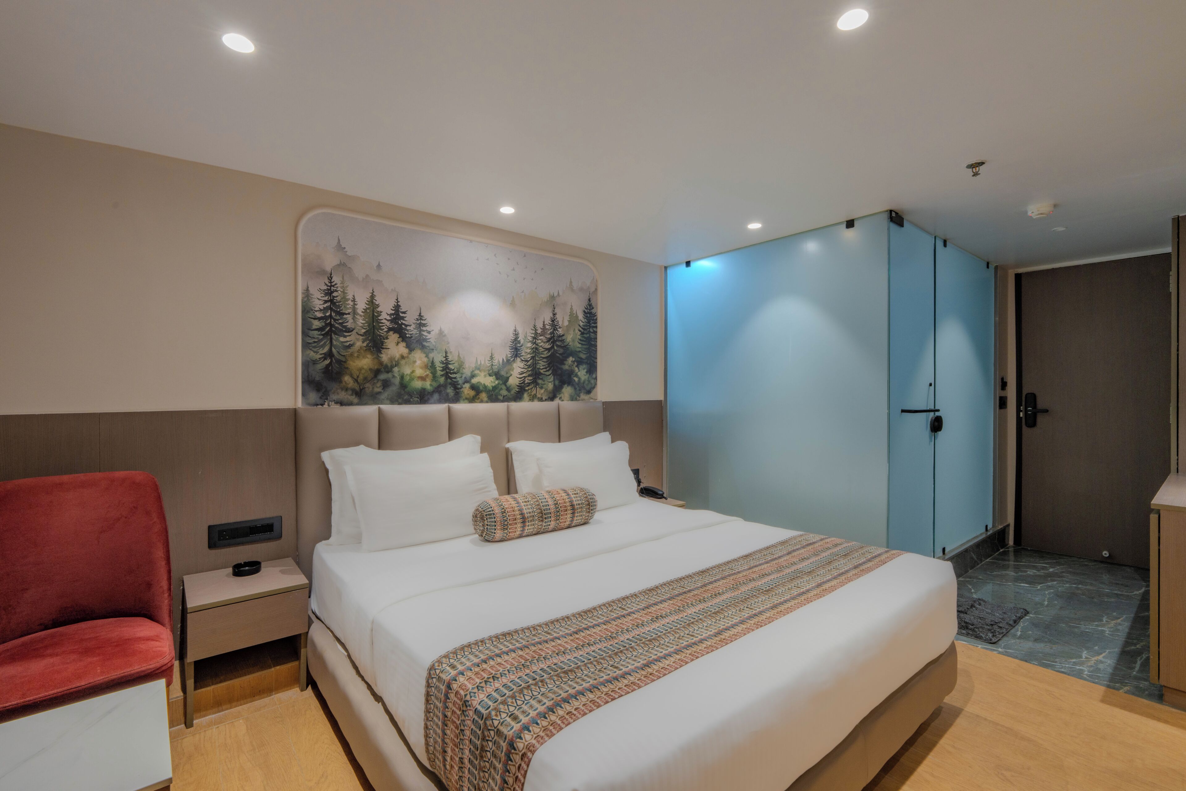 A modern hotel room with a large bed, a red chair, a wardrobe, and a painting of a forest on the wall.