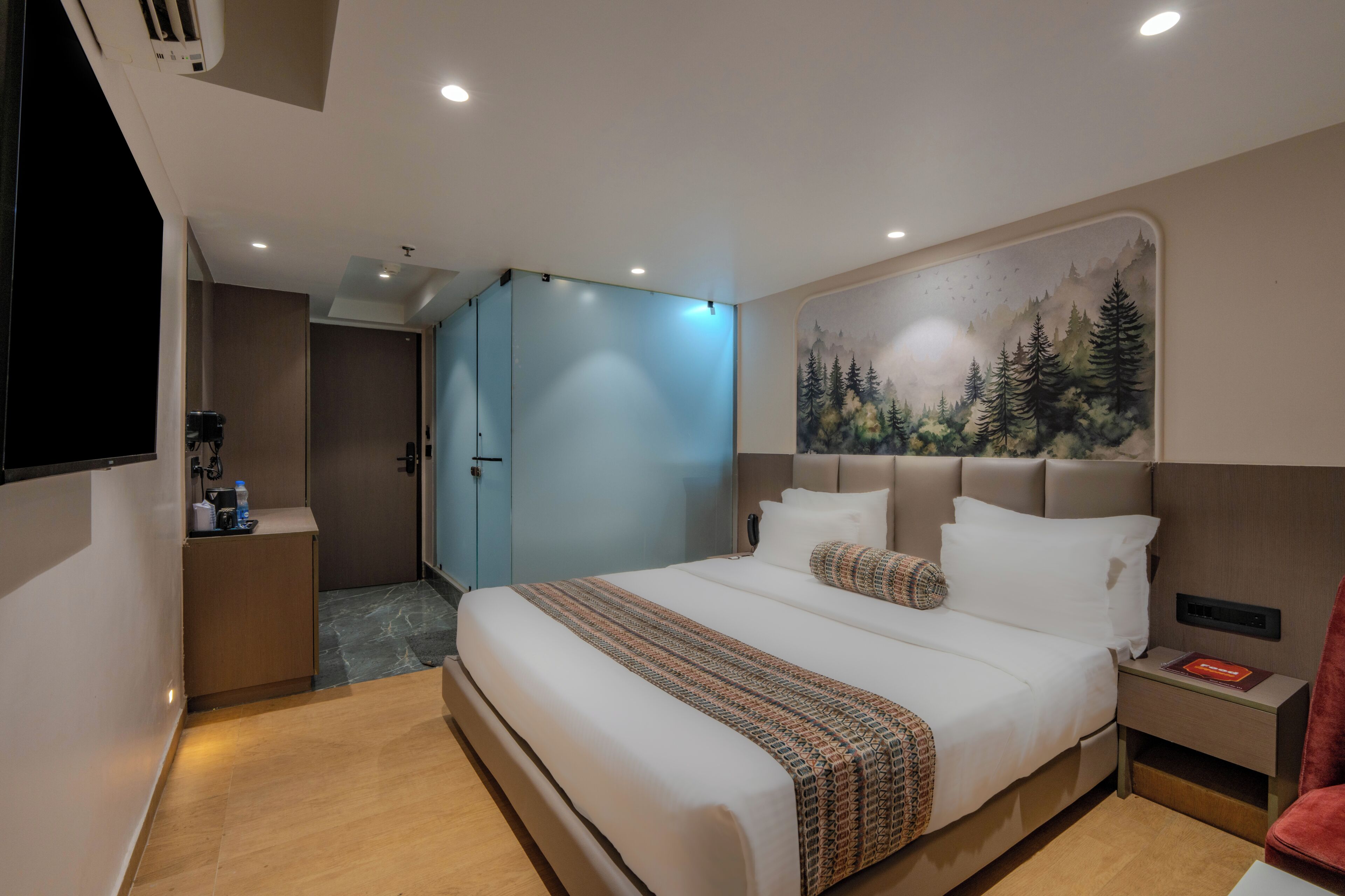A modern hotel room with a large bed, a headboard with a forest mural, a bedside table with a book, and a TV mounted on the wall.