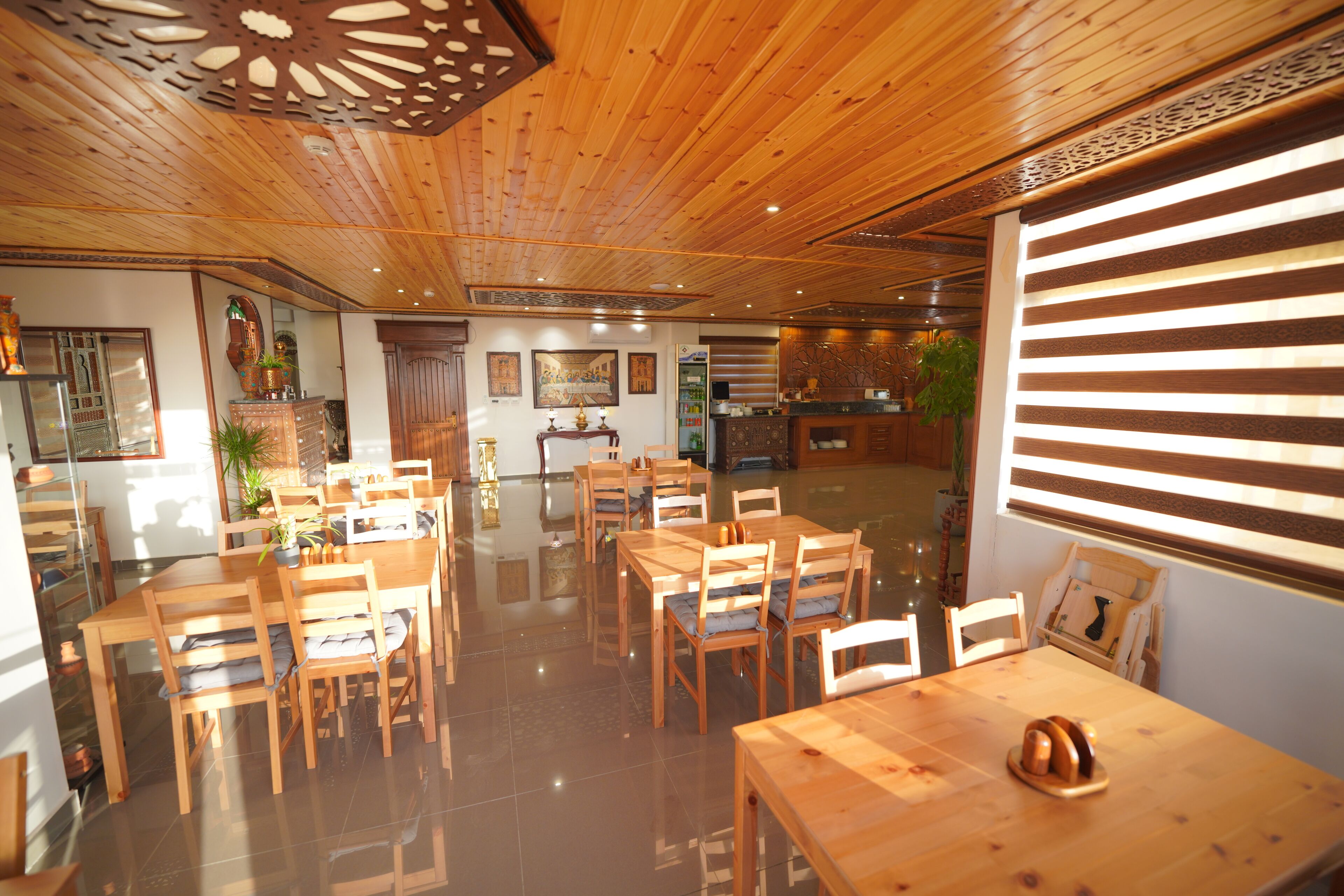 A wooden interior with multiple dining tables and chairs, a kitchen area, and a large window with blinds.