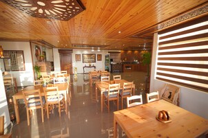 Un interior de madera con varias mesas y sillas de comedor, una zona de cocina y un amplio ventanal con persianas.