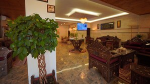A lobby with a large plant, wooden furniture, a chandelier, and a television on the wall. - Petra Plaza Hotel (Wadi Musa)