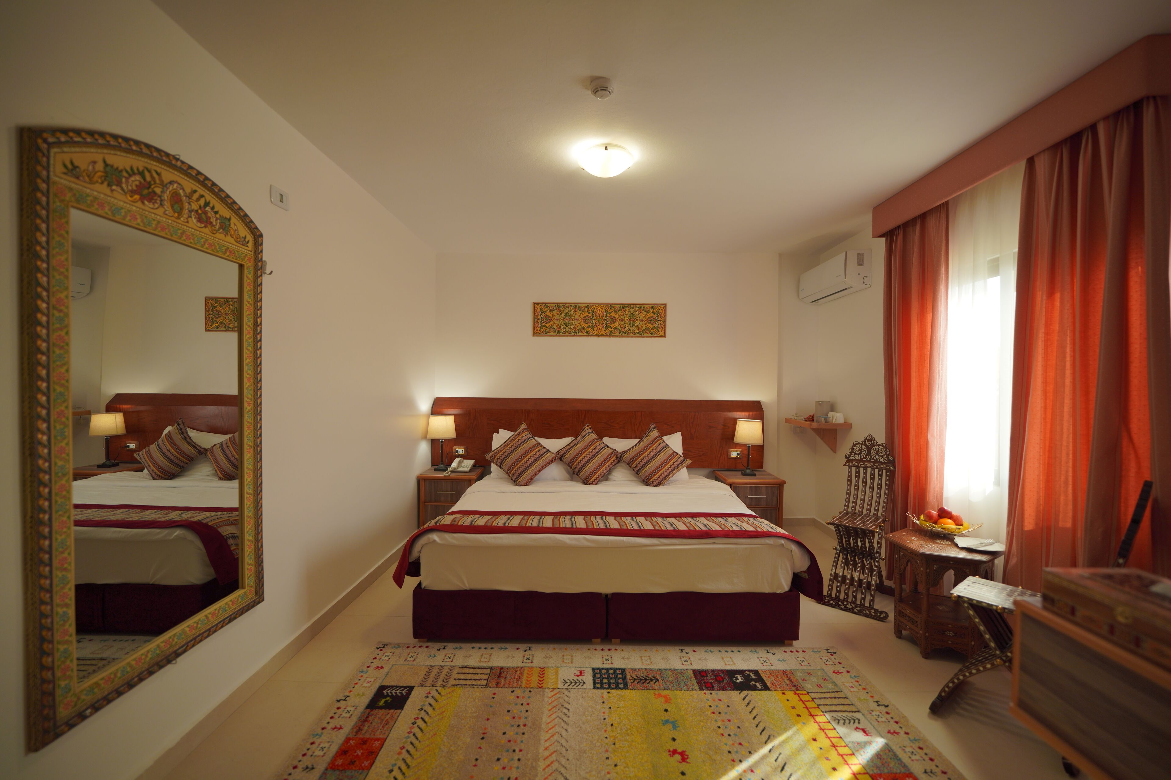 A hotel room with a large bed, a mirror, a headboard, a bedside table with a lamp, a small table with fruit, and a chair.