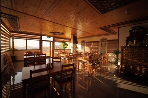 A spacious dining area with wooden tables and chairs, large windows with views of the mountains, and a decorative cabinet with plants. - Petra Plaza Hotel (Wadi Musa)