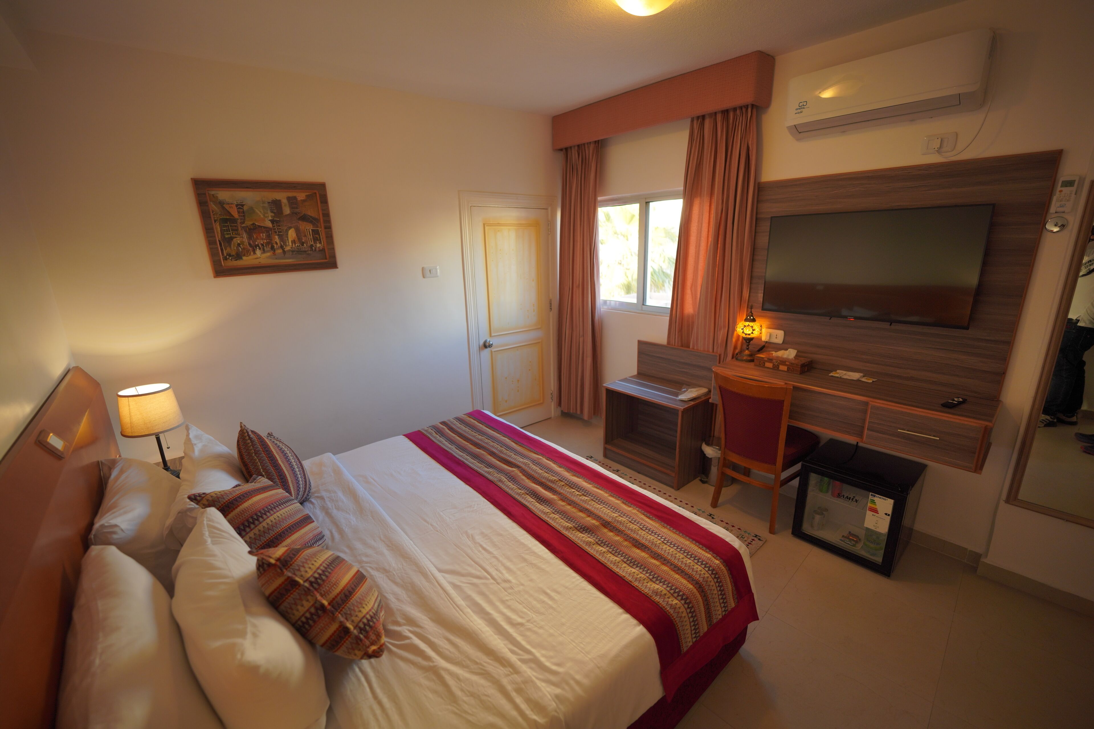A hotel room with a bed, a desk, a television, and an air conditioning unit.