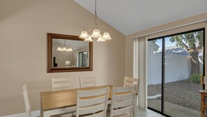 Un área de comedor con una mesa de madera y sillas blancas, un espejo grande y un ventanal corredero que da a un espacio exterior.