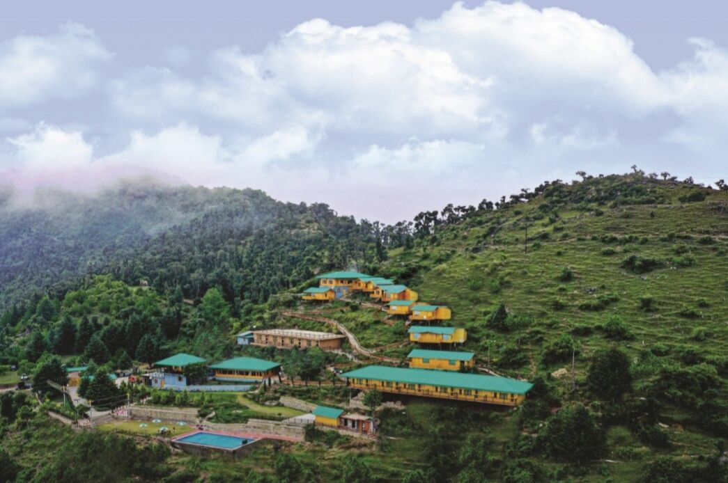 A cluster of yellow buildings with green roofs on a hillside, surrounded by lush greenery and trees.