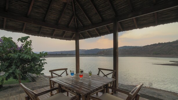 A wooden deck with a table and chairs overlooking a calm body of water, with a thatched roof overhead. - Toyam by Orchid Hotels (Bhor)