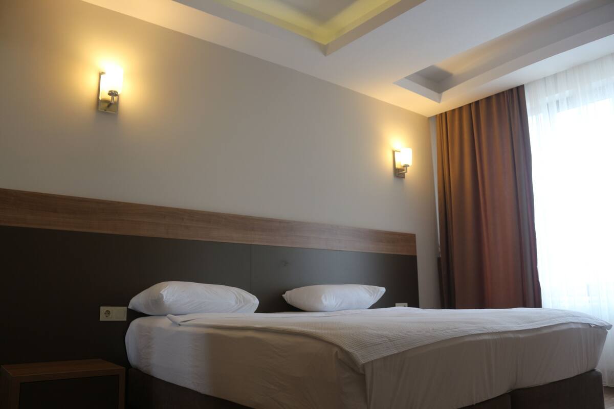 A hotel room with a bed, two wall-mounted lights, a window with curtains, and a wooden headboard.
