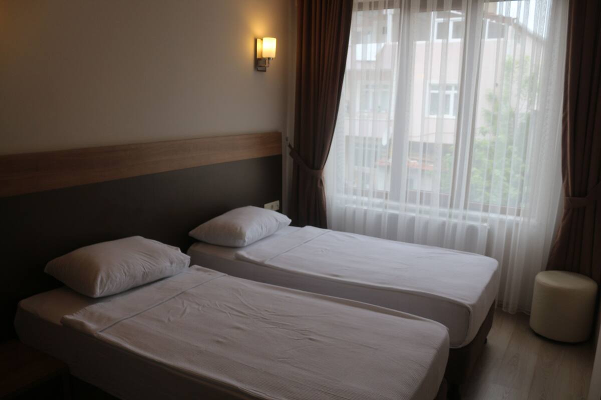 A hotel room with two single beds, a nightstand, a wall-mounted lamp, and a window with curtains.