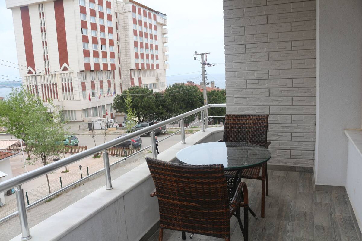 A balcony with wicker chairs and a glass table, overlooking a street with buildings and trees.