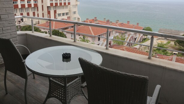 A balcony with a round glass table and wicker chairs, overlooking a coastal cityscape with red-roofed buildings and a view of the sea. - Longoz Hotel (Demirköy)