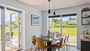 A modern dining area with a large table, chairs, and a view of a green landscape through large windows.