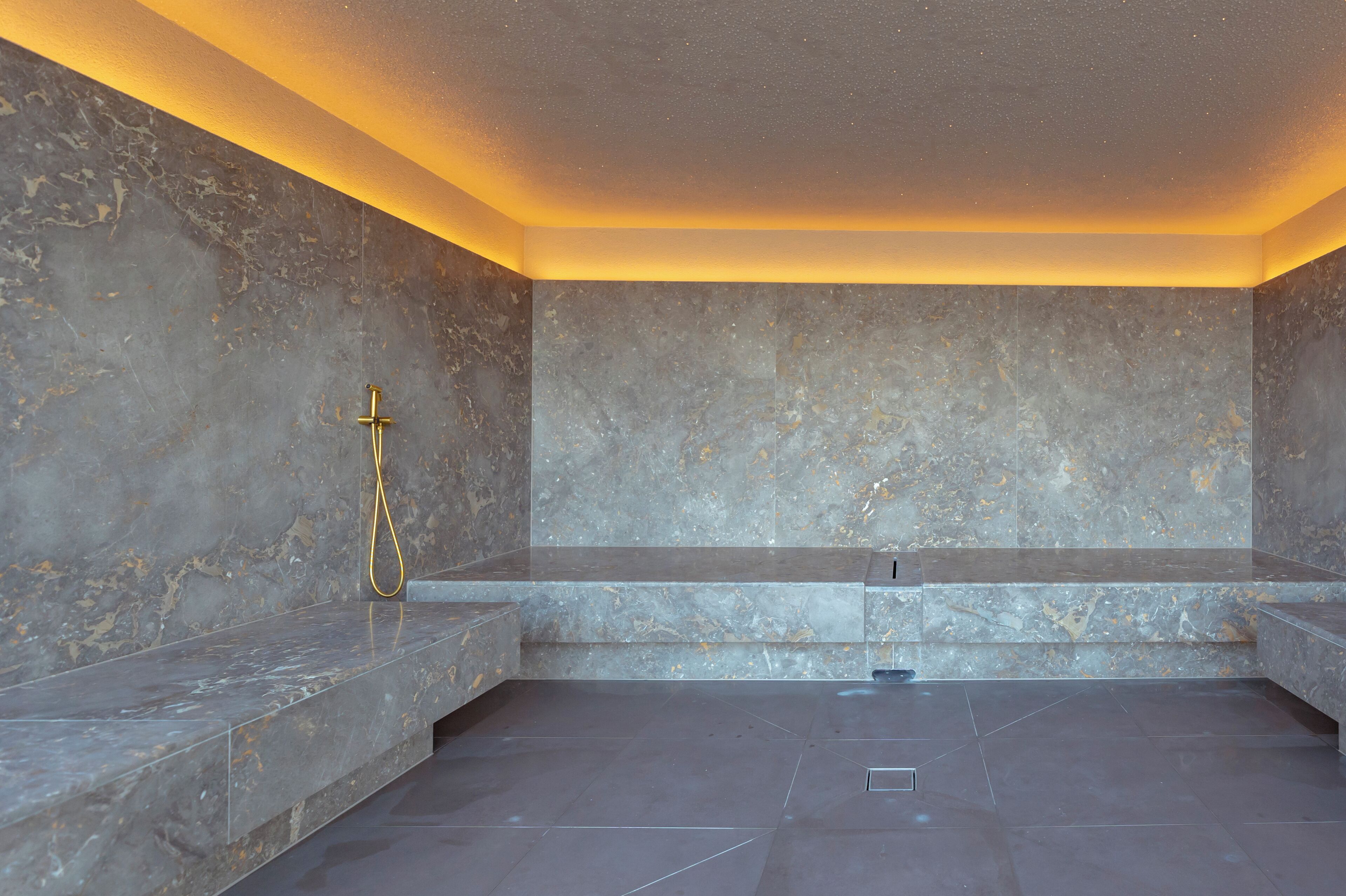 A modern bathroom with marble walls and floors, a gold showerhead, and a bench.