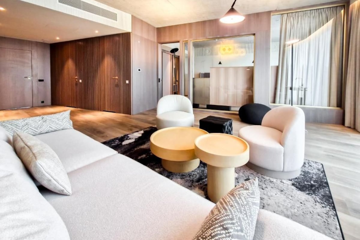 Una habitación de hotel moderna con una cama grande, dos sillones blancos, una mesita y un sofá.