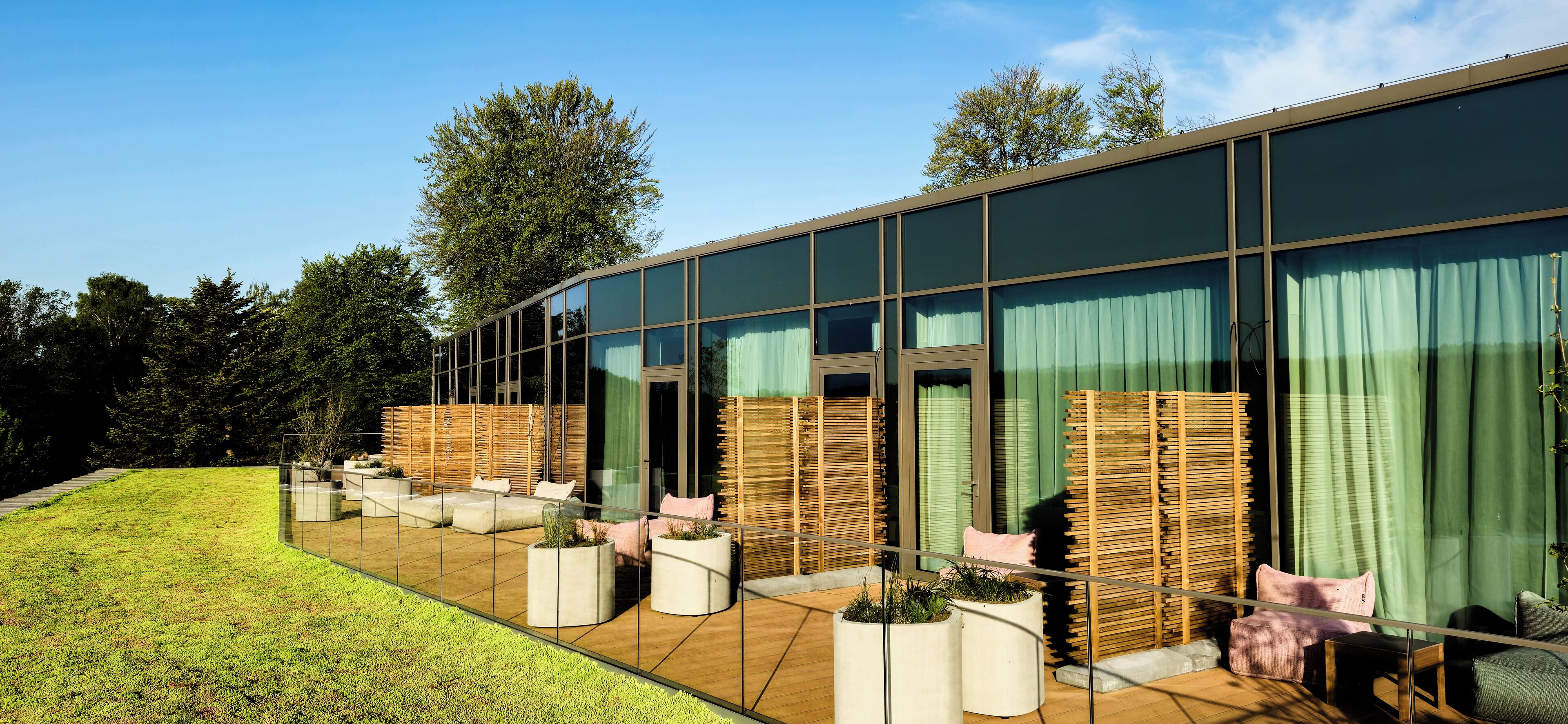 Een modern gebouw met grote glasramen, terraszitplaatsen met kussens en plantenbakken.