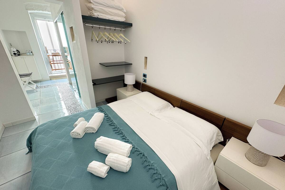 Een hotelkamer met een bed, nachtkastje, lamp, plank met handdoeken en uitzicht op buiten.