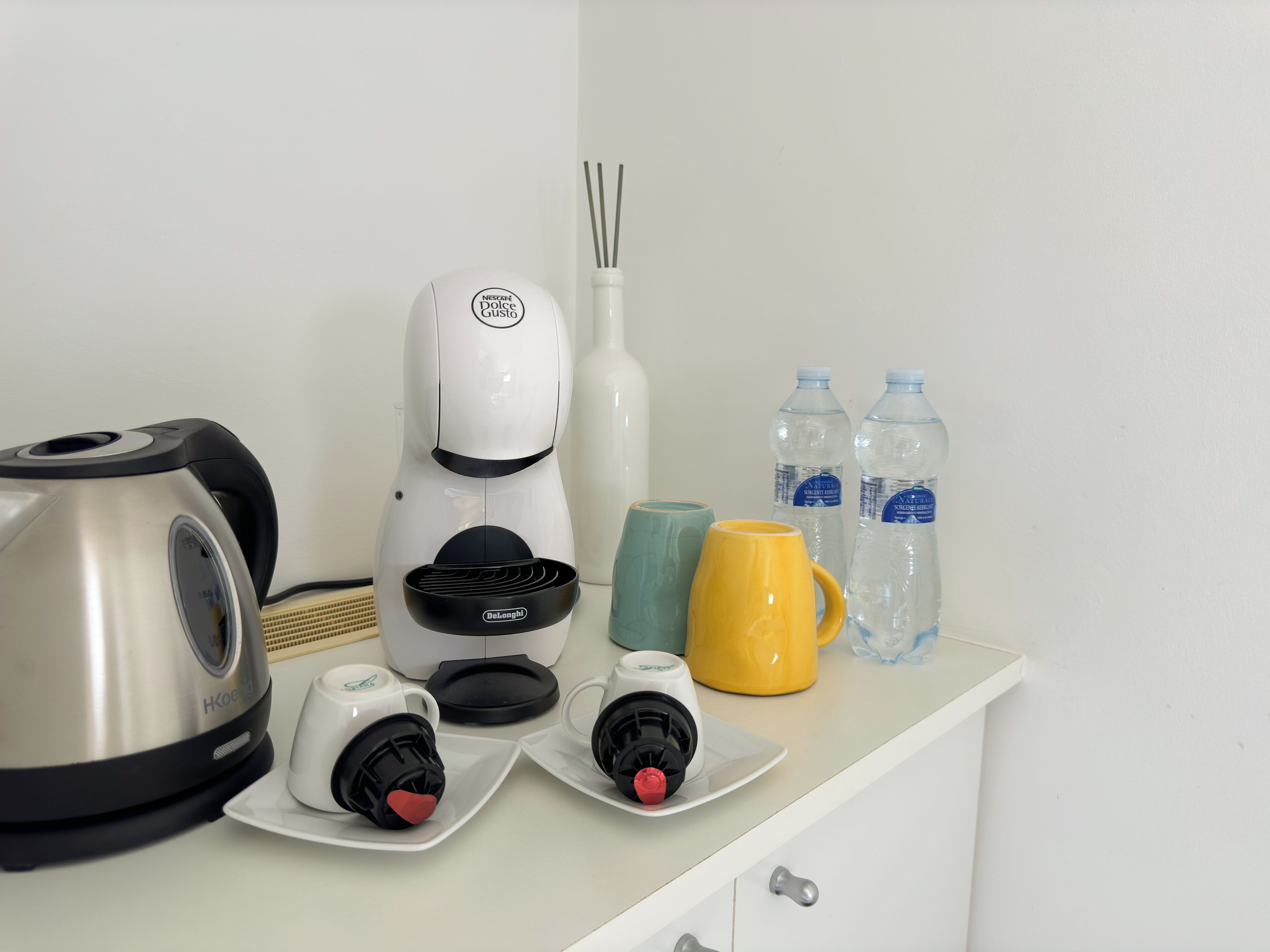 A kitchenette with a coffee maker, two mugs, a yellow mug, two water bottles, and a diffuser.