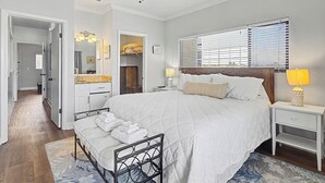 Ein Schlafzimmer mit einem Bett, Nachttischen, einem Fenster mit Jalousien und einem durchgehenden Türrahmen einsehbaren Badezimmer. Ein Deckenventilator ist ebenfalls vorhanden.