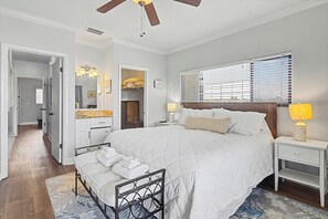 Un dormitorio con cama, mesitas de noche, un tendedero, ventilador de techo, ventana con persianas y un baño visible a través de una puerta abierta.