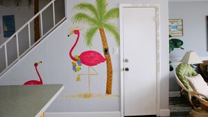 Ein Zimmer mit weißer Tür, einer Treppe, einem Wandbild mit Flamingo und Palme sowie einem Korbstuhl.