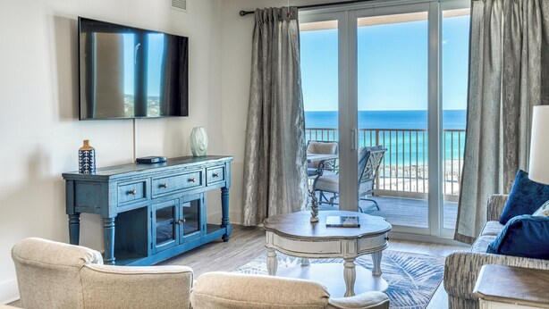 A room with a TV, a blue dresser, a round table, and a view of the ocean through a sliding glass door.