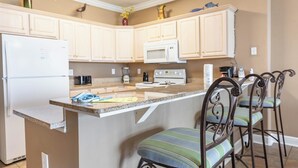A kitchen with white cabinets, a white refrigerator, a microwave, and a stove. There are two bar stools with striped cushions. - BIG Beachfront Balcony and New Furnishings (Panama City Beach)