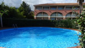 A swimming pool with a tiled edge, surrounded by greenery and a building with multiple balconies in the background. - la Castagna (San-Nicolao)