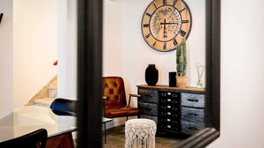 A room with a large wooden clock, a brown leather chair, a glass table, a black cabinet, and a small white stool.