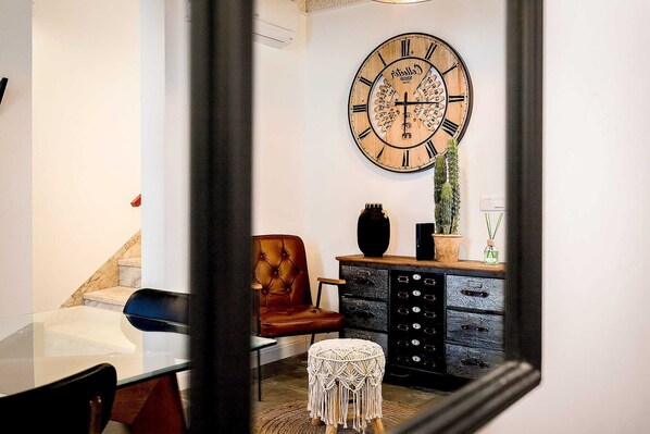 A room with a large wooden clock, a brown leather chair, a glass table, a black cabinet, and a small white stool.