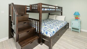 Un lit superposé avec une échelle en bois et des tiroirs, une petite table de chevet avec une lampe et une couvre-lit à motifs.