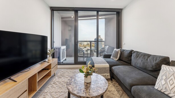 A modern living room with a flat-screen TV, a grey sofa, a coffee table with a vase, and a balcony with outdoor furniture. - Mega Style Apartments Kings Domain (Southbank)