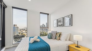 A modern hotel room with a large bed, a nightstand, and a view of the city through floor-to-ceiling windows. - Mega Style Apartments Kings Domain (Southbank)