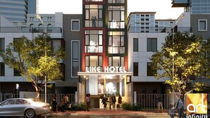 Un hôtel moderne, reconnaissable à son enseigne bien visible « Like Hotel », composé de plusieurs étages dotés de grandes fenêtres et d’une entrée centrale.