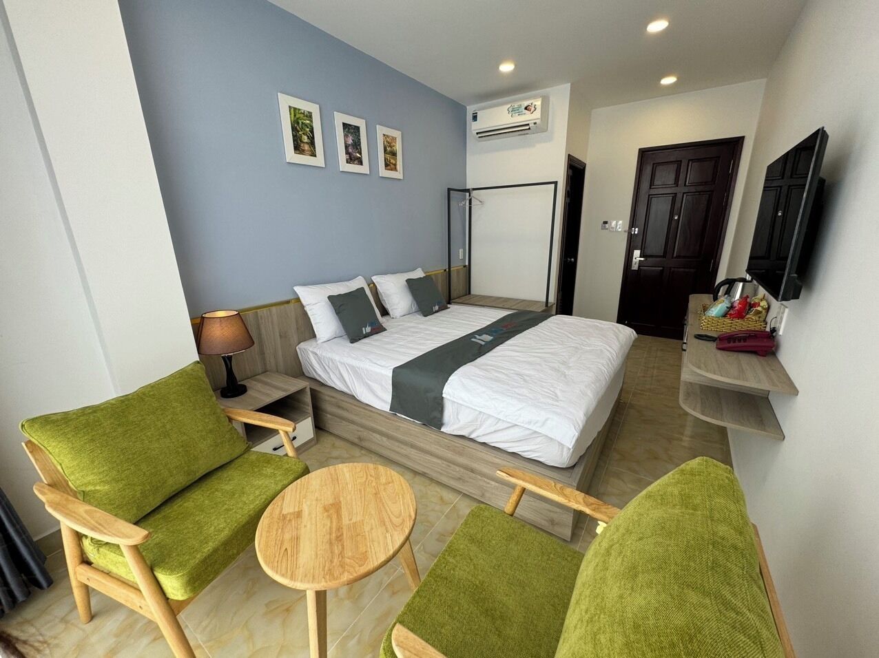 A hotel room with a bed, green upholstered chairs, a small wooden table, a TV mounted on the wall, and a door.